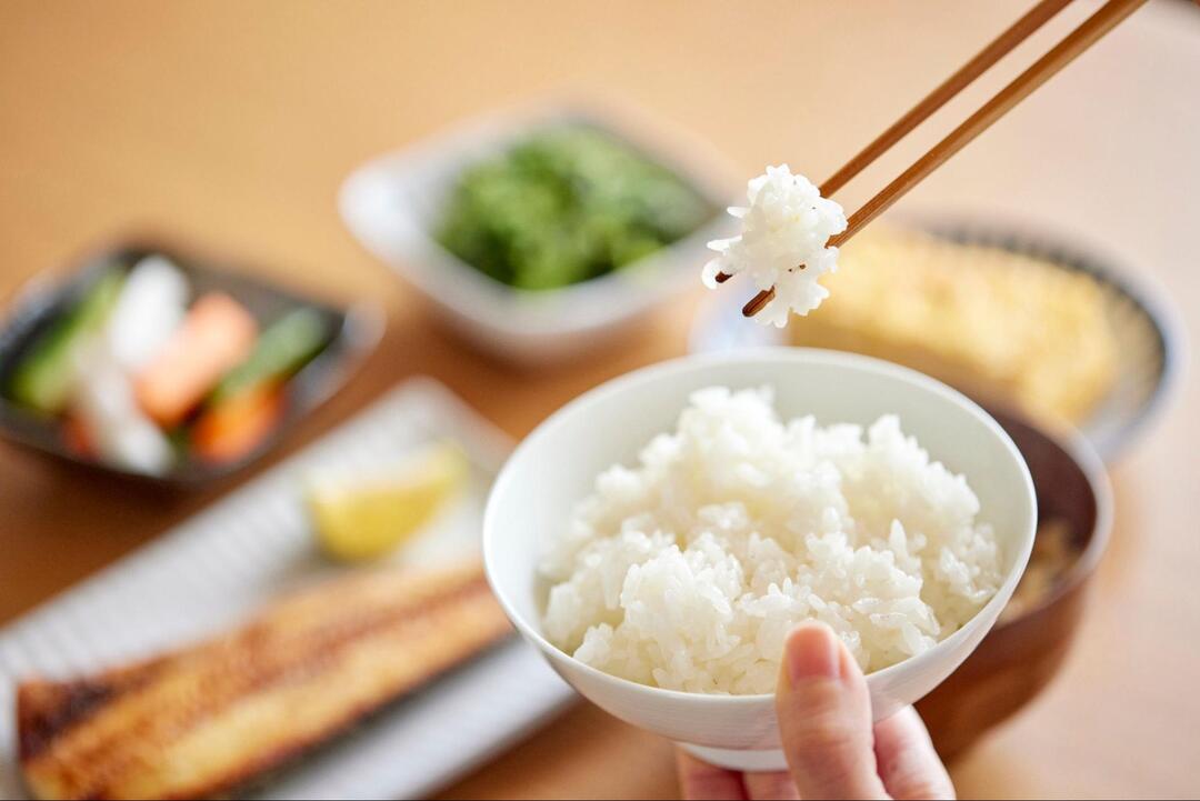 箸で白米をひとくち分持ち上げているお茶碗の手元のアップ。背景には焼き魚や小鉢などの和食が並んでいる。