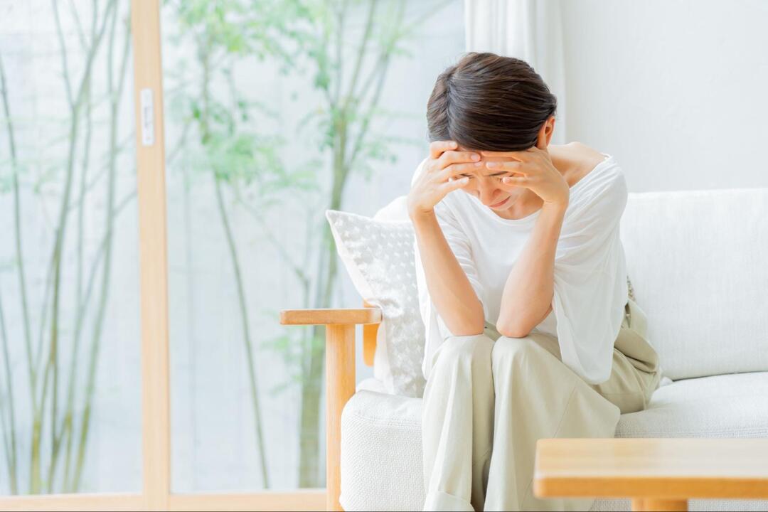 ソファーに腰掛けた女性が、両手を額に当てて下を向き、体調不良や痛みに耐えている様子