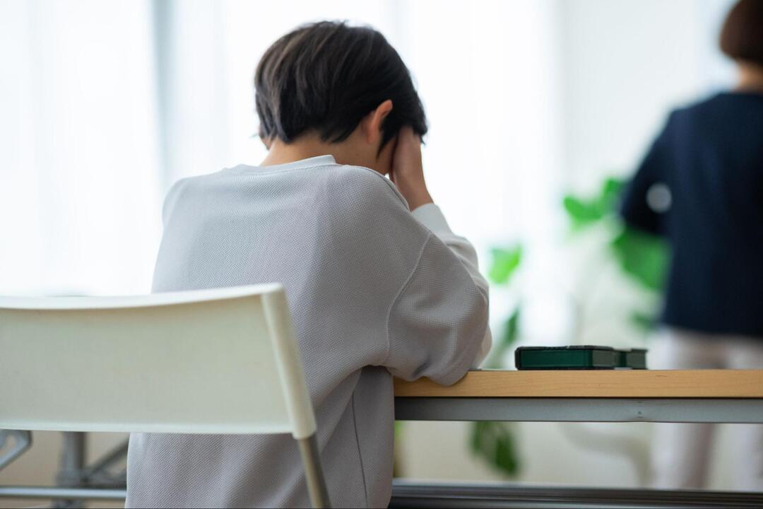 机の前に座る短髪の人物が、後ろ向きの状態で両手で顔を覆い、うつむいて悩んでいる様子