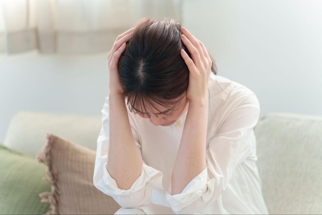 女性がソファーに座り、両手で頭を抱え込むようにして深くうつむいている様子