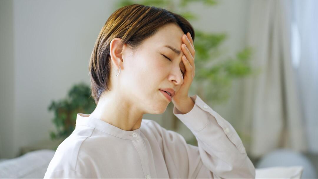 女性が目を閉じながら左手で額を抑え、頭痛や体調不良に耐えている様子