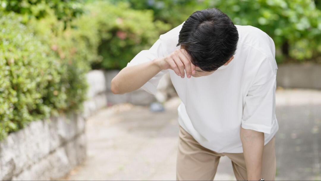 男性が屋外で前かがみになり、右手で額を抑えながらめまいや体調不良に耐えている様子