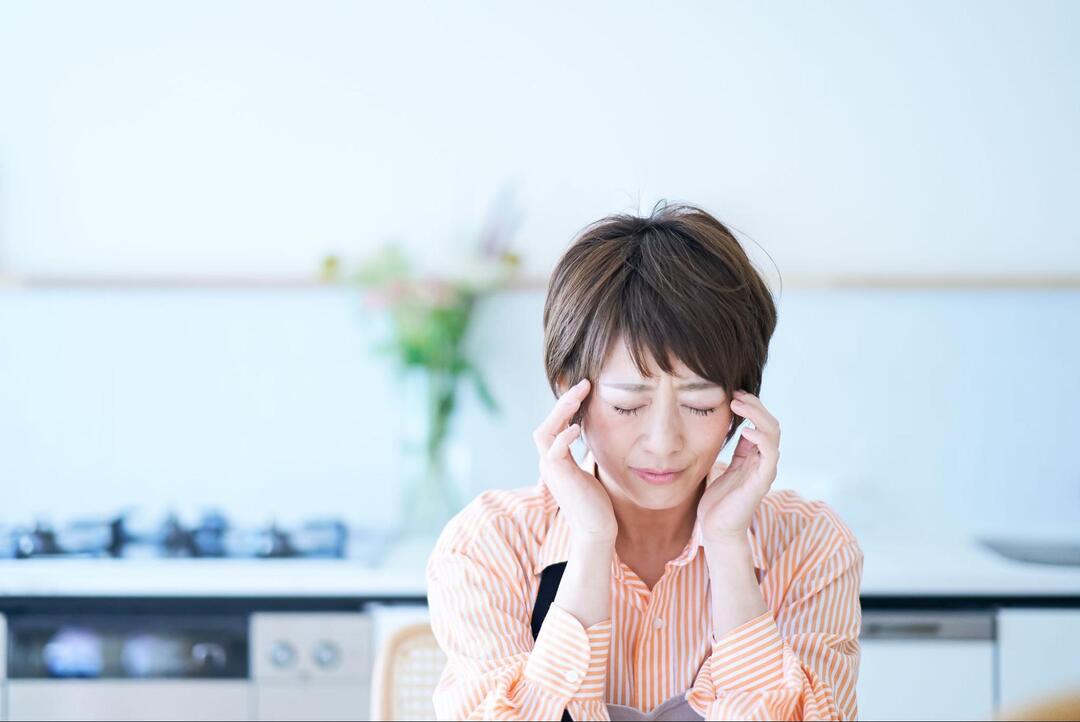 女性が目を閉じて眉間にシワを寄せ、両手でこめかみを押さえて頭痛に耐えている様子