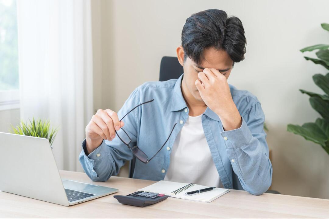 デスクの前でメガネを外し、目頭を押さえながら目が疲れた様子でうつむいているシャツ姿の男性