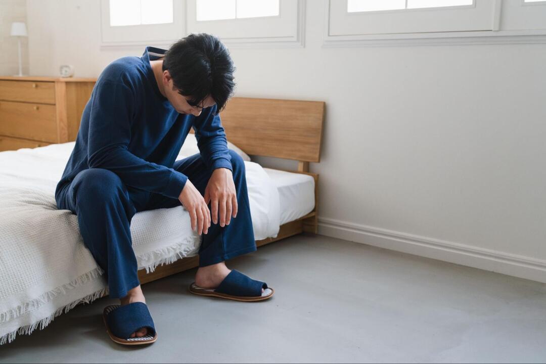 紺色のパジャマを着た男性がベッドの縁に腰掛け、力なく下を向いてうなだれている様子