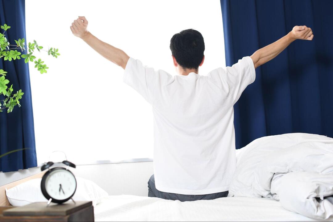 白いTシャツを着た男性がベッドの上に座り、両腕を大きく広げて背伸びをしている後ろ姿