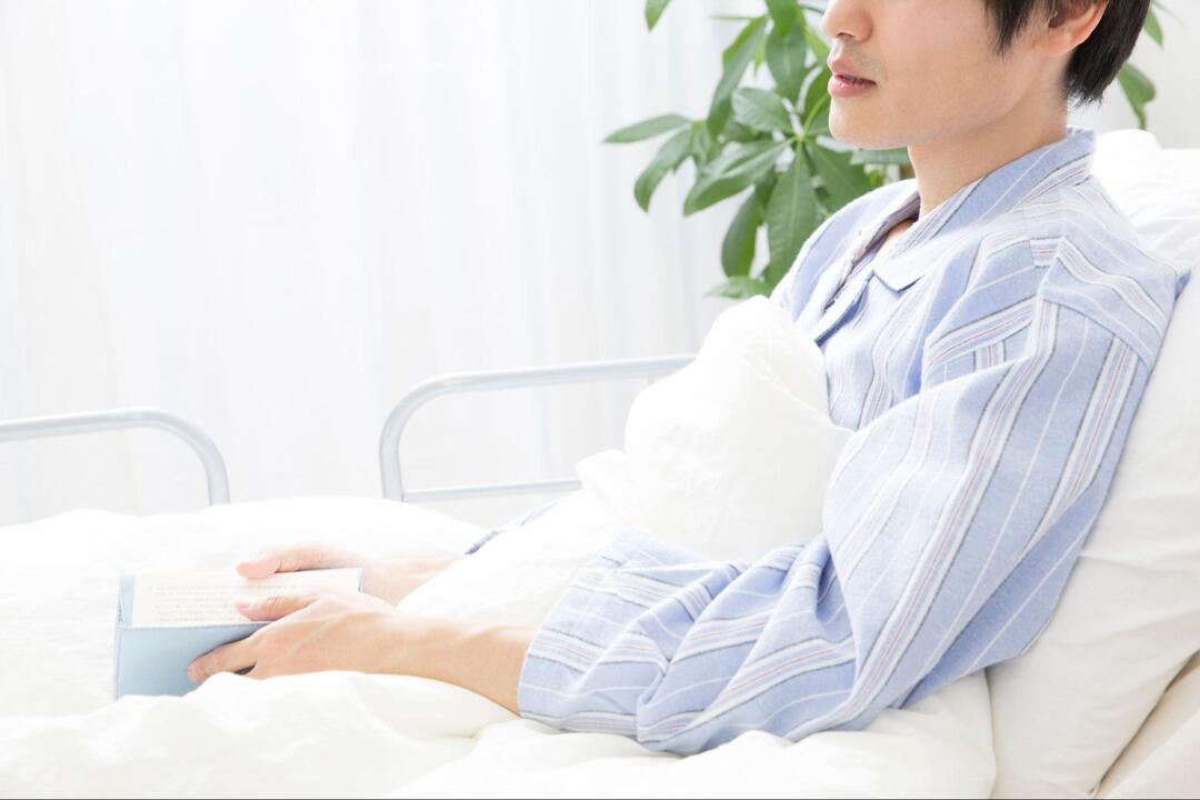 病院のベッドに上体を起こして座り、本を手に持っている男性