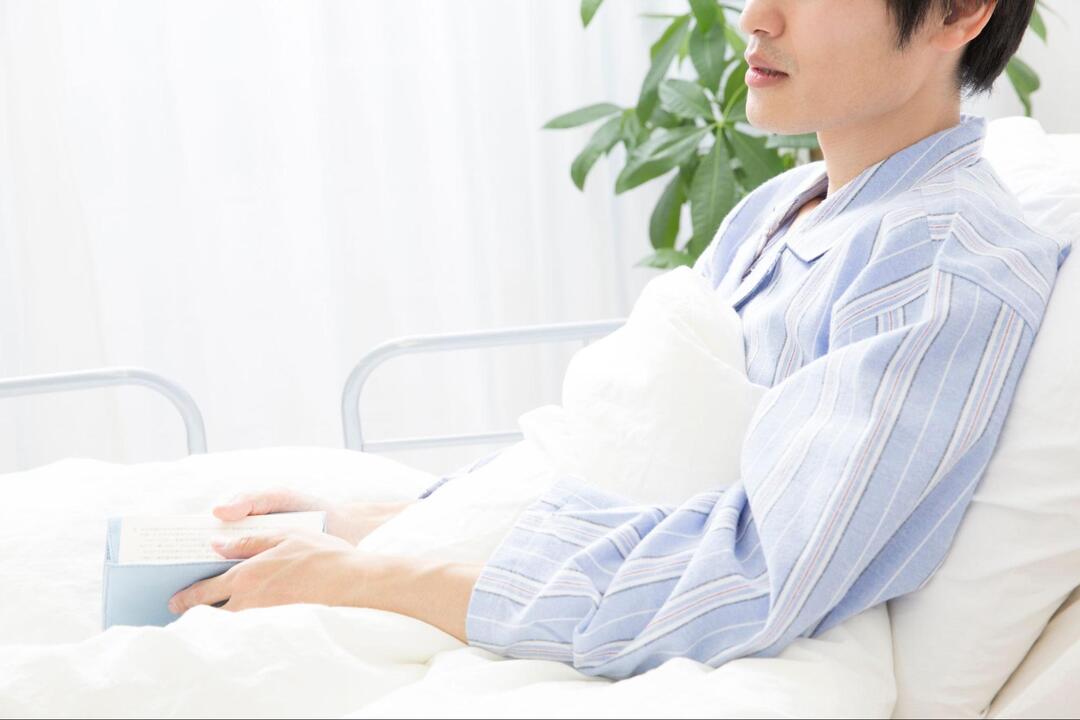 病院のベッドに上体を起こして座りながら本を手に持っている男性